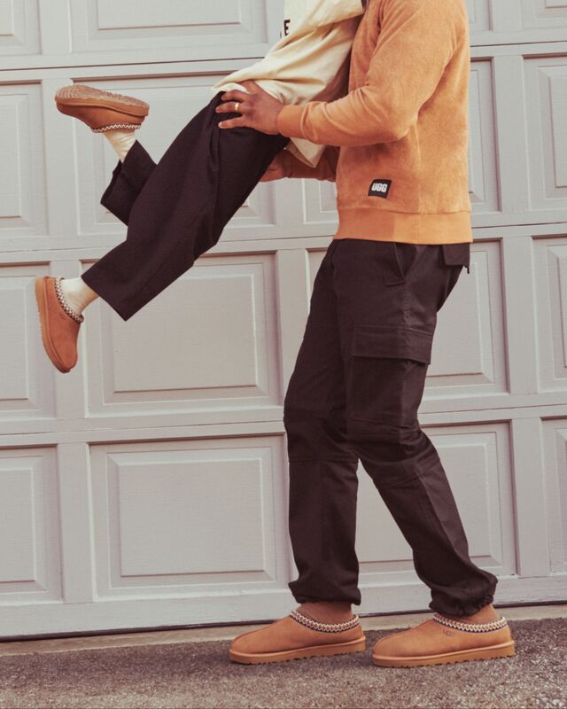 A photo of a man wearing a Kamryn Crewneck sweatshirt and Tasman slipper lifting a child wearing a Tasman II Slipper for children.