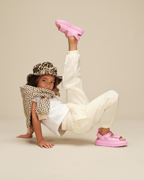 Un enfant tout en beige et blanc, faisant une pose acrobactique, portant des chaussures UGG roses.