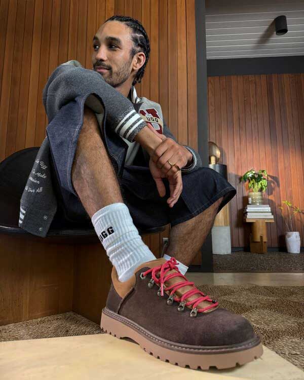A masculine person seated in a well designed, retro style room wearing a stylish fit and the new UGG Birch Hiker.