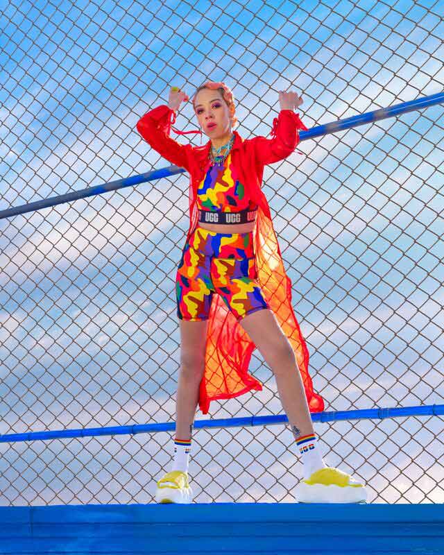 A femme presenting person wearing UGG Pride gear, leaning against a chain link fence.