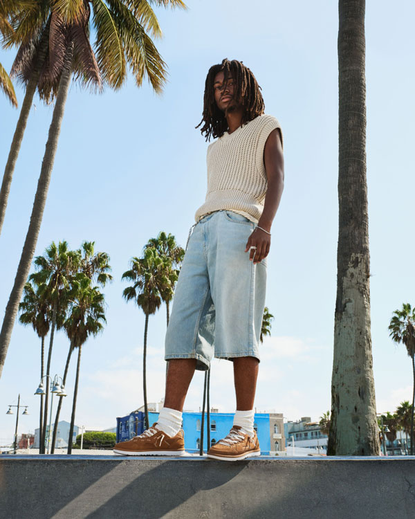 Un homme debout à l’extérieur sur un mur de béton, portant des chaussures UGG, avec des palmiers et un ciel bleu.