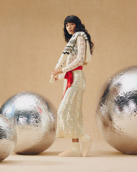 A feminine person framed by playfully oversized metallic orbs, wearing UGG slippers and a matching fit accessorized with a festive scarf.