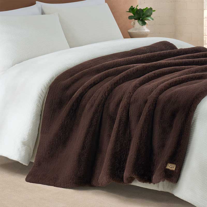 A plush throw blanket in an earth tone color draped over a neatly made bed.
