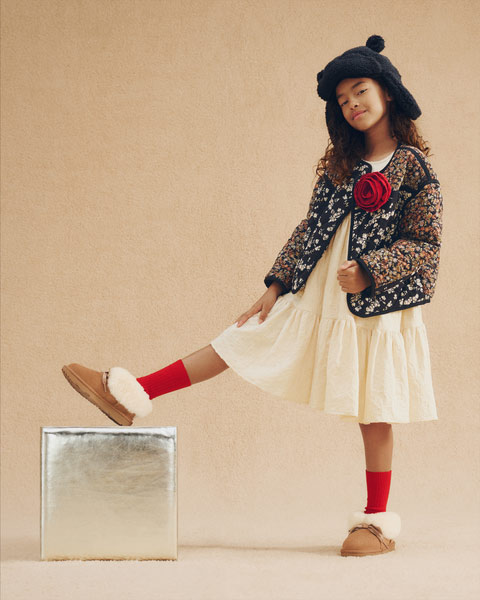 A kid showing off their fluffy UGG slipper, also wearing a fun and festive fit and a big, cozy winter hat.