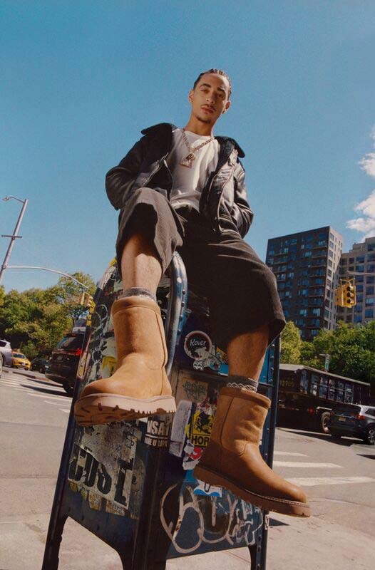 Une personne masculine assise sur une boîte aux lettres couverte de graffitis et d’autocollants, portant une coupe assortie et la botte UGG Birch II de retour.